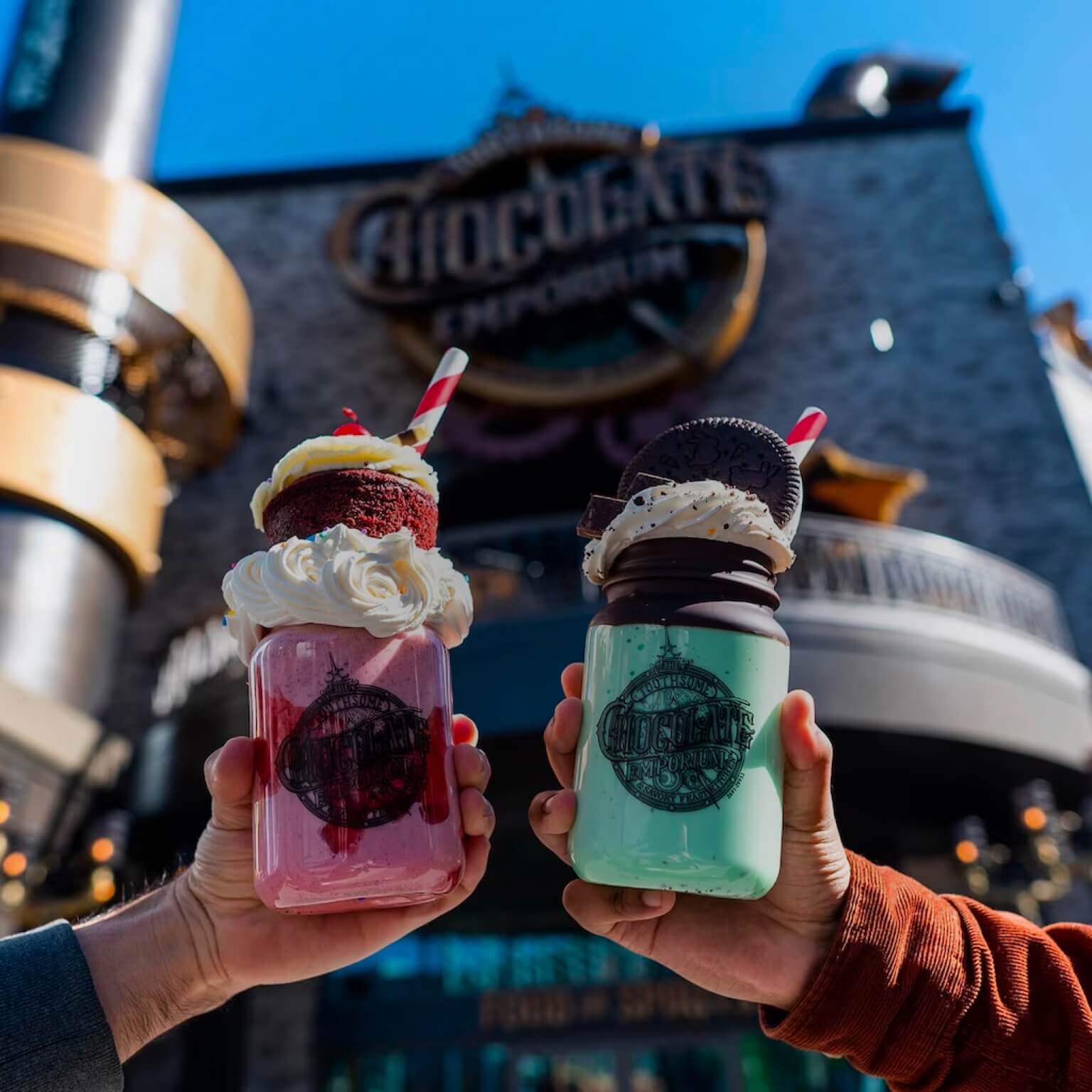 Two hands holding milkshakes in front of the exterior entrance sign to The Toothsome Chocolate Emporium & Savory Feast Kitchen.