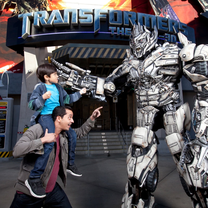 A dad with his son on his shoulders pointing at a transformer in front of the Transformer ride.