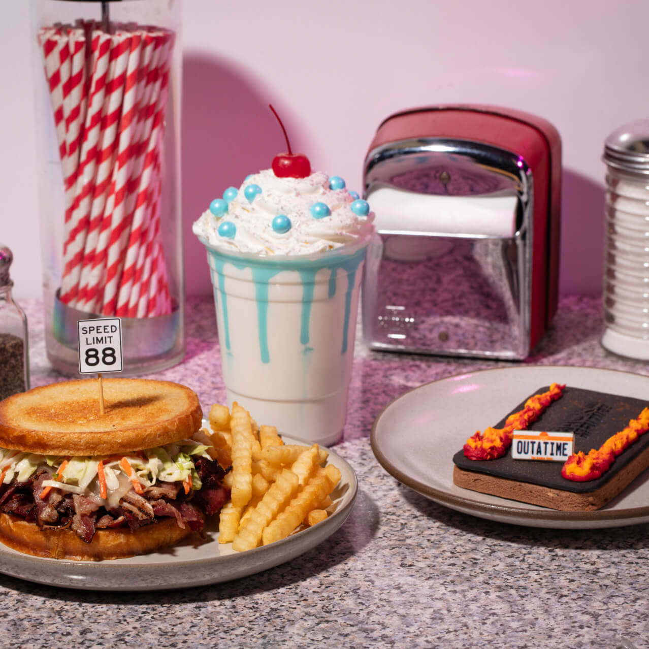 A diner style table setting presents a meal with a Back to the Future theme, featuring a brisket sandwich with fries, a white and blue milkshake, and a rectangle cookie with “OUTATIME” lettering on top.