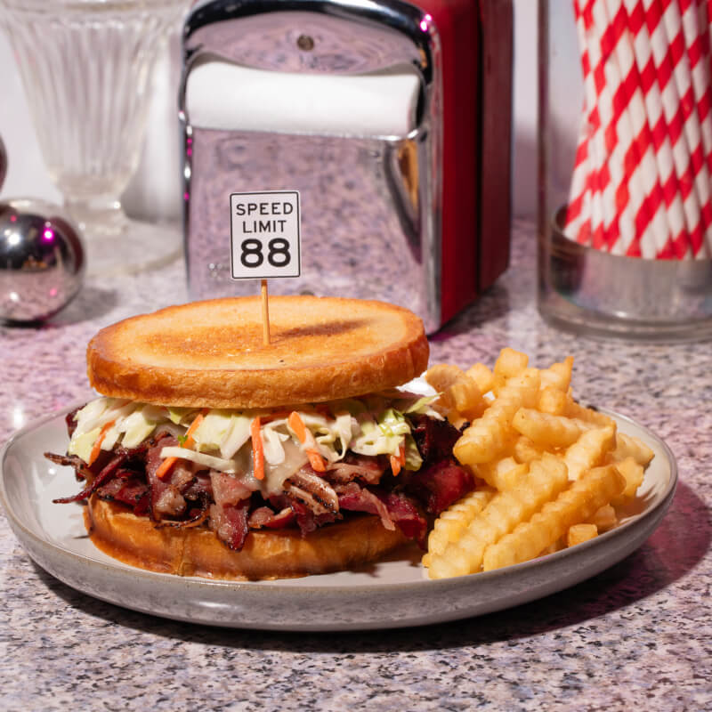 A brisket and coleslaw sandwich with a small flag on top of the bread that reads “SPEED LIMIT 88” next to a side of fries.