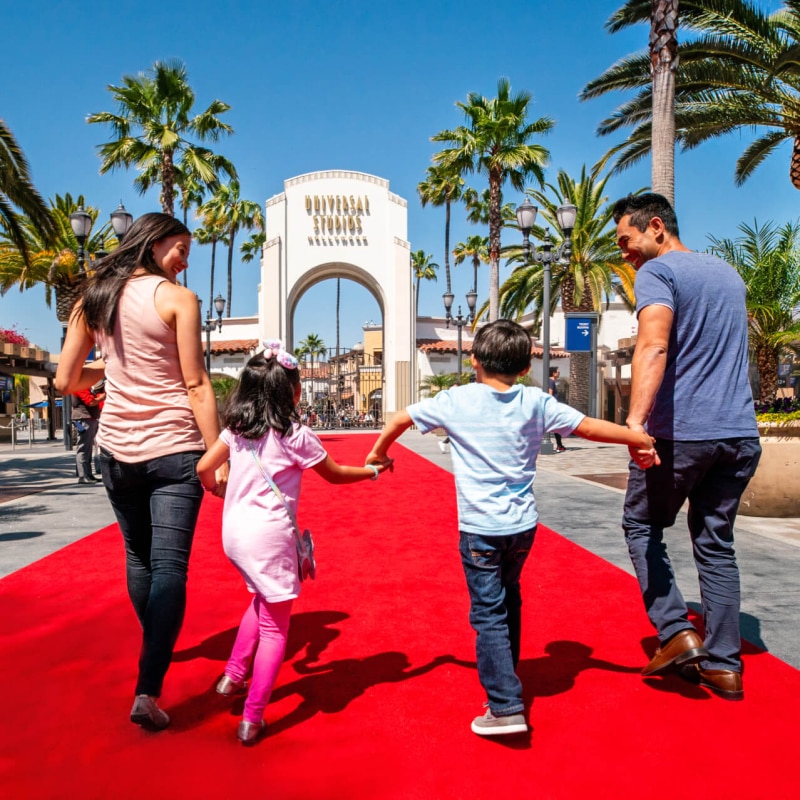 A family standing on a red carpet walks happily towards the arch entrance of Universal Studios Hollywood.