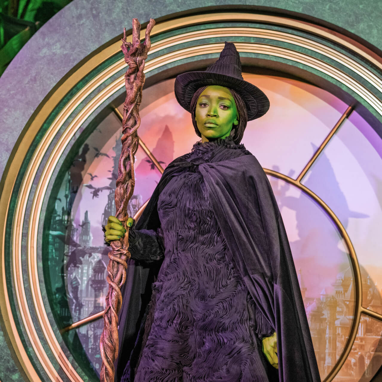 Serious Elphaba character in full black costume holding her broom and posing in front of a round pink-and-green Oz photo backdrop.