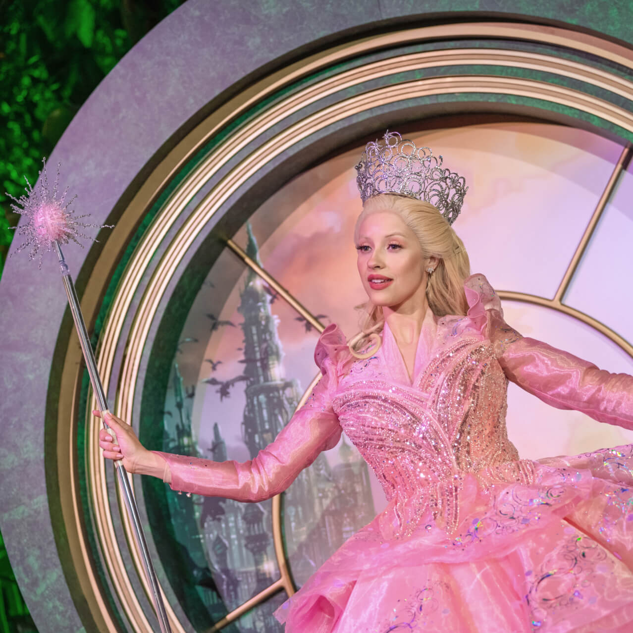 Smiling Glinda character in a pink dress and crown holding her wand and posing in front of a round pink-and-green Oz photo backdrop.