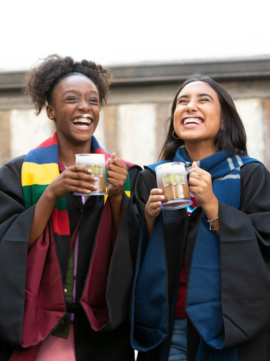 A pair of young women wearing Hogwarts gowns and holding mugs of Butterbeer smile and show off their white Butterbeer mustaches.